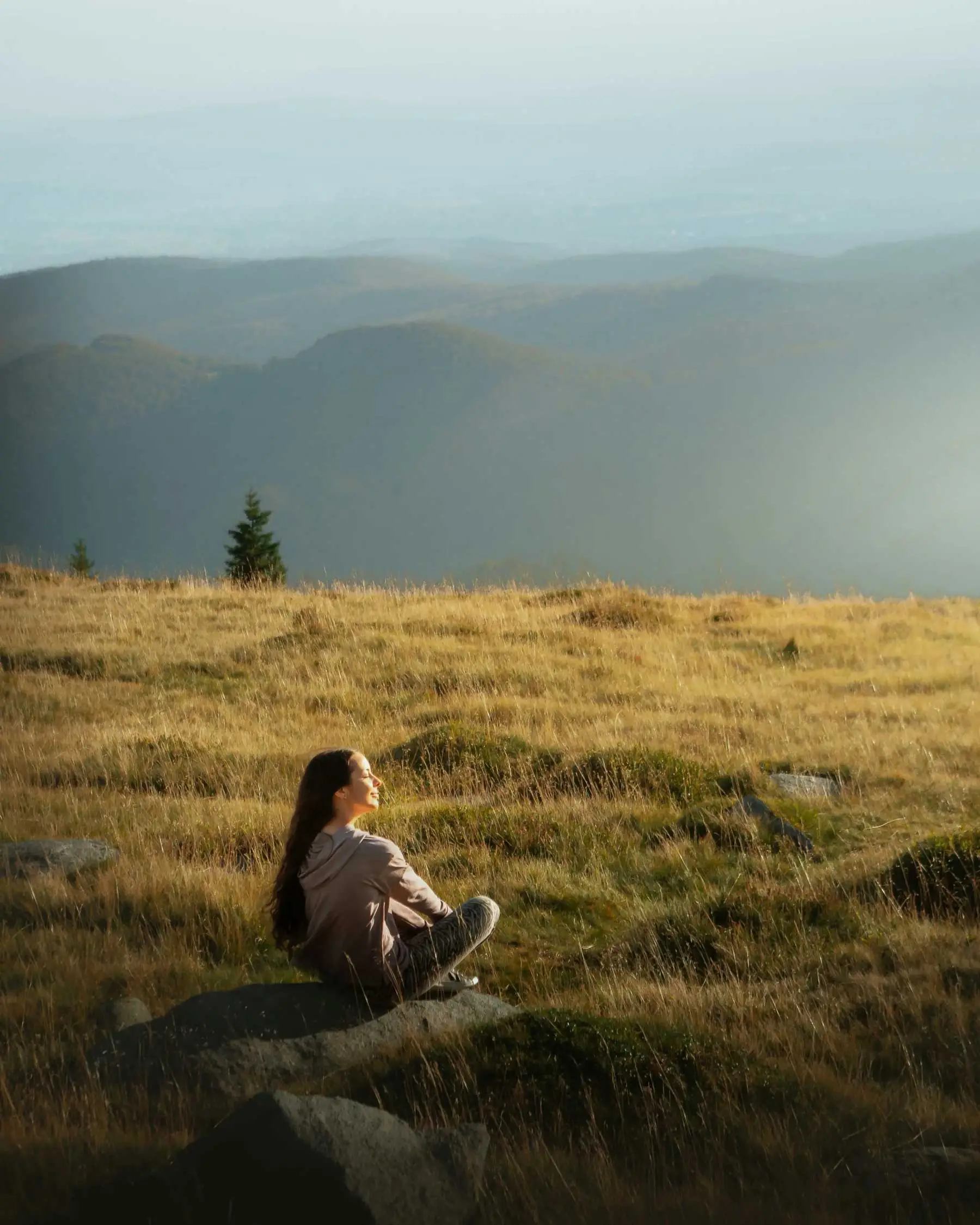 Une femme détendue, assise seule dans un pré en pleine nature, face au soleil.